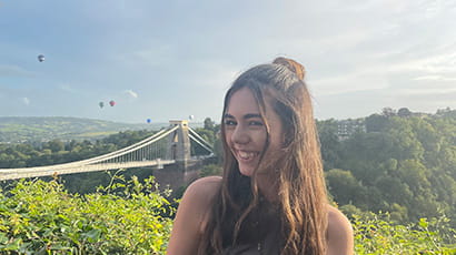 UWE Bristol student Emily Annakin stands smiling with the Clifton Suspension Bridge behind her with hot air balloons flying in the distance