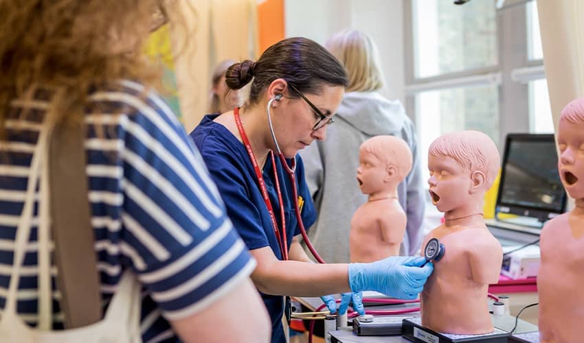A medical clinician demostrates uses a stethescope on a child manequin while a young adult watches on.