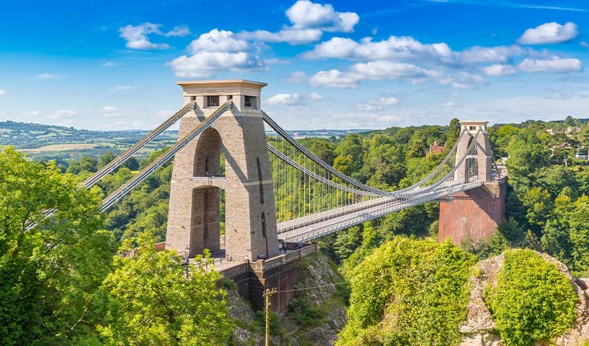 The image shows the Clifton Suspension Bridge, a famous suspension bridge spanning the Avon Gorge and the River Avon in Bristol, England. The bridge features two large stone towers with cables extending from them to support the roadway. The surrounding area is lush with greenery and trees, and there are hills in the background under a partly cloudy sky.