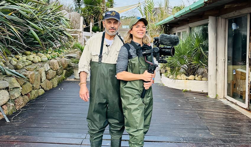 Two individuals wearing green waders stand on a wooden pathway outdoors. The person on the right is holding a camera with a large microphone attached. They are surrounded by lush greenery and stone walls, with buildings visible in the background.