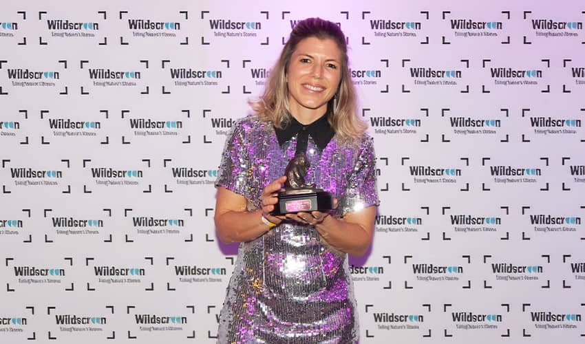A woman wearing a shiny dress smiles at the camera, while holding an award