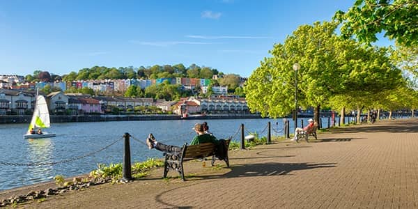 A sunny day at Bristol Docks with a young couple sitting on a bench with a row of colourful houses beyond the water.