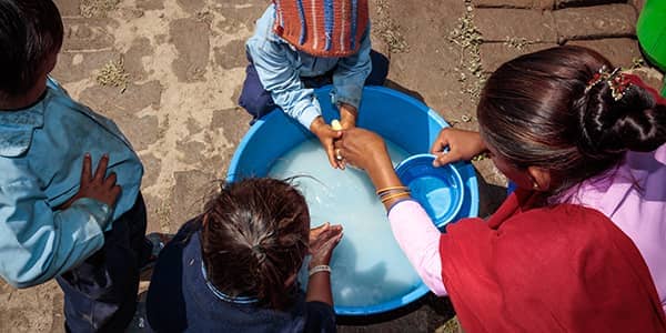 A female helps children wash their hands in a bucket of water.