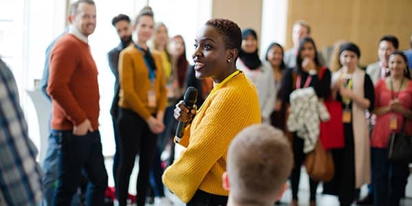 Smiling black female with a microphone in hand addressing a smiling standing crowd of mixed races.