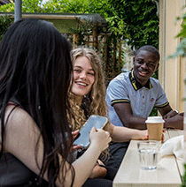Three students sat together laughing at a window bench outside a cafe.