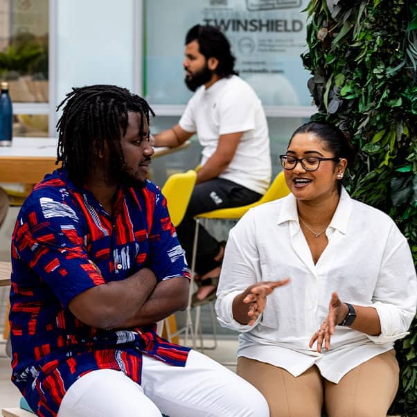 Man and woman in smart-casual dress sit talking animatedly next to a large indoor plant in an airy office space.