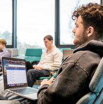 A student sits on a chair with a laptop on their lap.