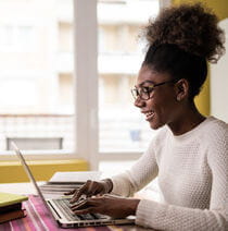 A woman smiling and sitting at a table typing on a laptop