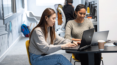 Two students working on laptops together at a small desk in the UWE Bristol Library.