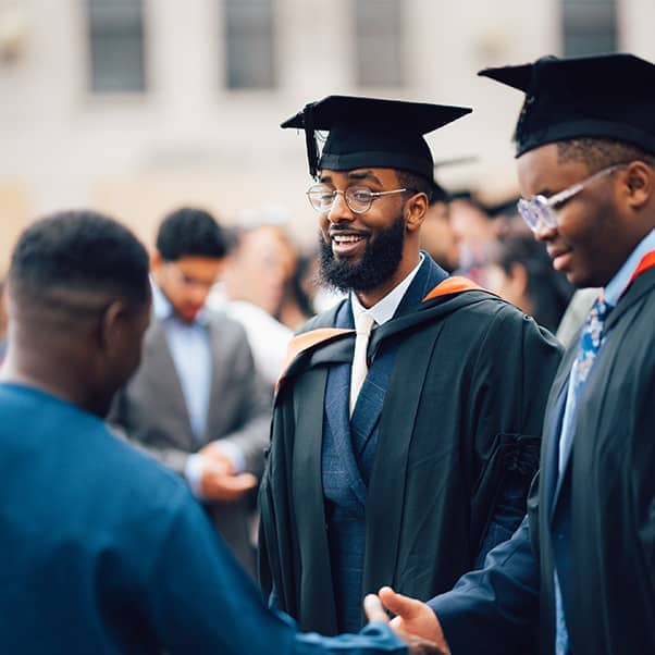 Two students in full graduation gowns standing together, one shaking hand of a friend in front.