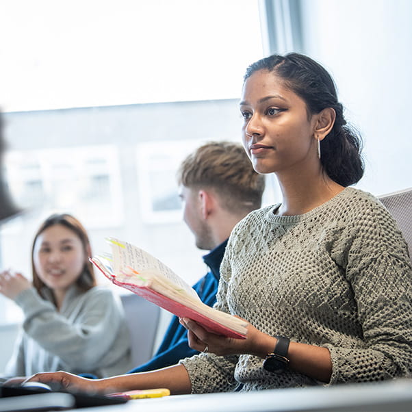 A student looking at a computer screen in the library with two students talking behind her